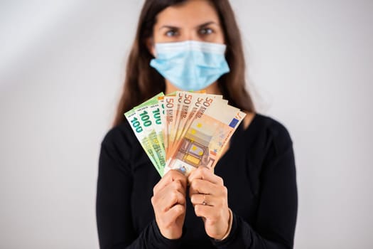 Happy woman holding multiple banknotes and wearing face mask in studio. Portrait of cheerful female adult with long brown hair with money in her hands.