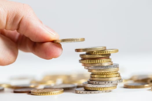 Holding coin in fingers over the stack of money from close up. Adding one euro on pile of metalic cash. Concept of saving salary for better profit.