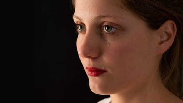 Sad woman crying on black background from close up. Unhappy girl with tears falling from eye on dark backdrop. Younge female feeling upset.