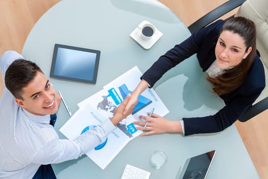 Top view of young Business partners closing a deal.Couple shaking hands over deal with documents and digital tablet on table showing statistics and graphics.