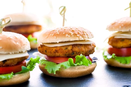 Macro close up of Multiple mini chicken burgers. Spicy chicken filets with cheese and tomato with sesame seed buns.