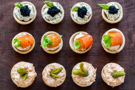 Close up Top view of variety of multiple mini puff pastry tartlets on wooden table. Open pastries filled with seafood.