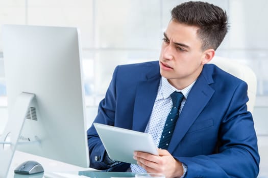 Close up portrait of young Businessman synchronizing digital tablet with computer. Young man sharing information between computer and digital tablet at desk in office.