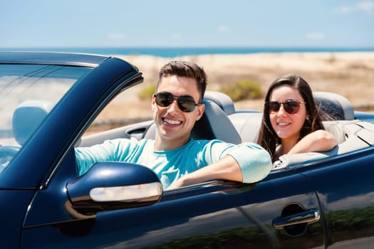 Close up portrait of handsome young man driving convertible along seaside with girlfriend sitting in back.