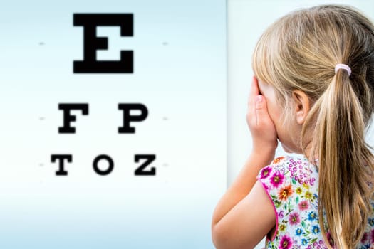 Close up rear view of girl testing eyesight. Infant closing one eye with hand looking at vision test chart.