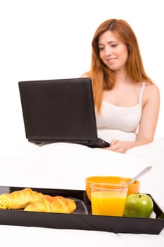 Young beautiful woman having breakfast in bed while working or relaxing with her computer