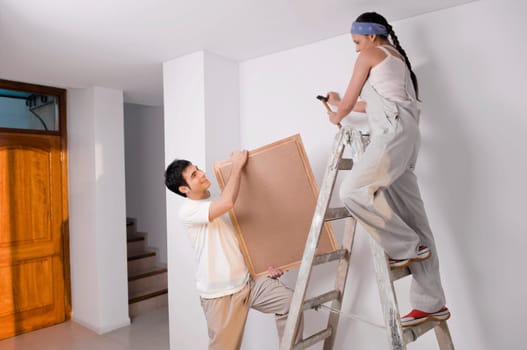 Young woman hammering nail on the wall with a young man holding a picture frame