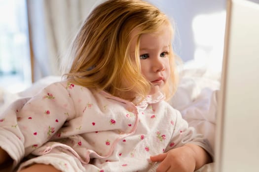 Close_up of a baby girl leaning on the bed and looking at a laptop