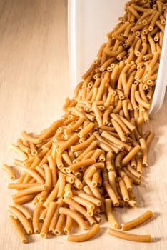 Wholewheat macaroni spilling out of a plastic container which has been placed on it's side on a wooden surface.