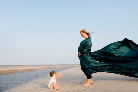 Side profile of a pregnant woman standing on the beach with her baby girl sitting in front of her