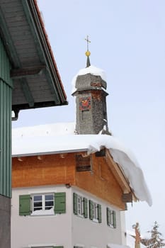 A dangerous roof avalanche on a house roof in front of a church in winter. Snowy church tower in Bavaria
