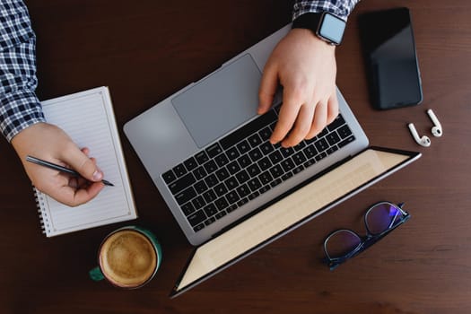 Man working at the laptop with a cup of coffee, phone, writing with pen at home. High quality photo