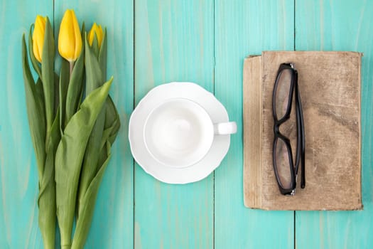 Old book with glasses,yellow tulips and empty cup on wooden background