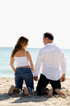 Rear view of a mid adult man and a young woman kneeling with holding hands on the beach