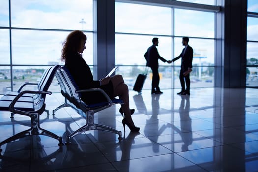 Young woman with touchpad waiting for departure on background of handshaking business partners