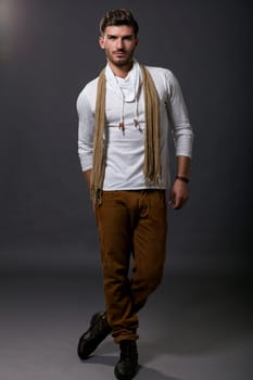 Stylish young man posing in smart casual clothes with a scarf jumps casually in front of the camera against a grey background
