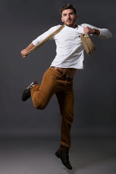 Stylish young man posing in smart casual clothes with a scarf jumps casually in front of the camera against a grey background