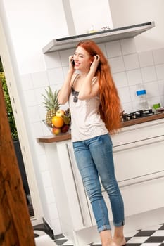 Happy young woman with mobile phone smartphone at home in the kitchen while making phone calls with long red hair