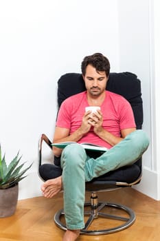 Young man in casual clothes relaxes while reading in an armchair with a drink in his hand
