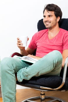 Young man in casual clothes relaxes while reading in an armchair with a drink in his hand