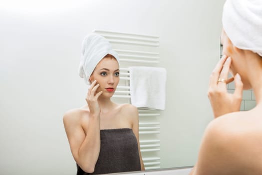 Young woman wrapped in a towel looks in the mirror in the morning in the bathroom