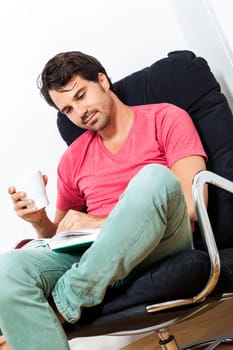 Young man in casual clothes relaxes while reading in an armchair with a drink in his hand