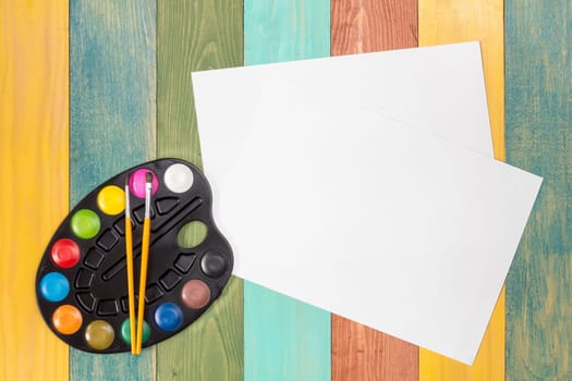 Watercolor paint and white paper sheet on color wooden background. Top view