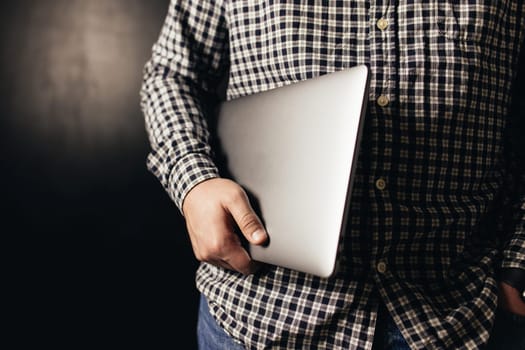 Man wearing casual clothes holds closed laptop, black blurred background. High quality photo