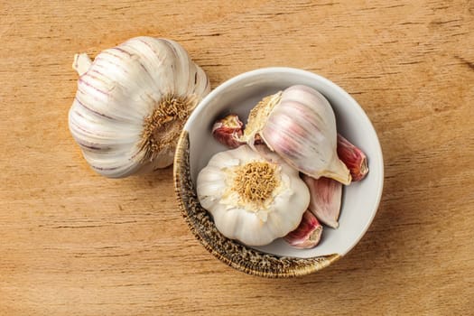 Table top view on garlic bulbs and cloves in small ceramic bowl, placed on old wooden cutting board.