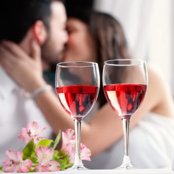 Close up of two wineglasses with rose wine and flower bouquet.In background a out of focus couple kissing.