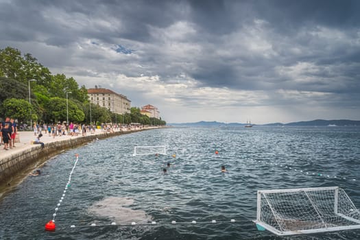 Zadar, Croatia, July 2019 Crowds of tourists walking and relaxing on the Coast of King Petar Kresimir IV. People playing water polo in Adriatic Sea