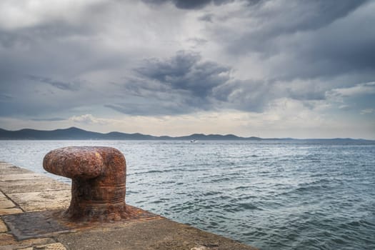 Single, old and rusty mooring bollard on the pier with a view on Adriatic Sea and dramatic sky in Zadar bay, Croatia
