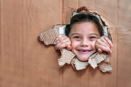 Smiling girl behind a cardboard, looking through a large hole in the cardboard and with her hands on the edge of the hole