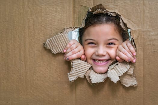 Smiling girl behind a cardboard, looking through a large hole in the cardboard and with her hands on the edge of the hole