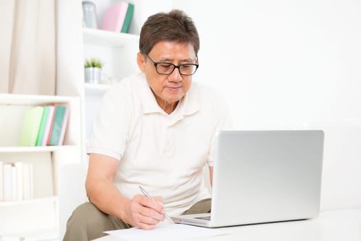 Portrait of mature Asian man using computer laptop and writing something on paper, sitting on sofa at home, senior retiree indoors living lifestyle.