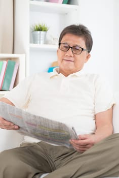 Portrait of mature Asian man reading on newspaper, sitting on sofa at home, senior retiree indoors living lifestyle.