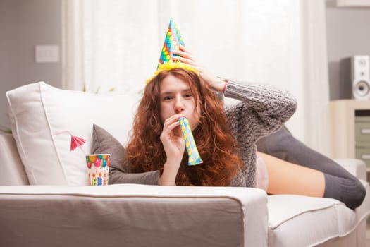 Red Headed very cute girl smiling on her sofa in her living room having some fun in aparty with a carton cone hat, a drink and party-props