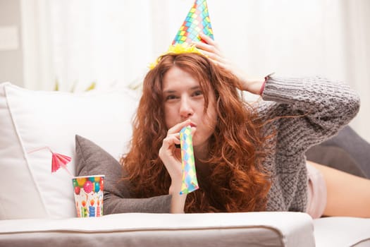 Red Headed very cute girl smiling on her sofa in her living room having some fun in aparty with a carton cone hat, a drink and party-props