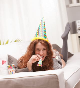 Red Headed very cute girl smiling on her sofa in her living room having some fun in aparty with a carton cone hat, a drink and party-props