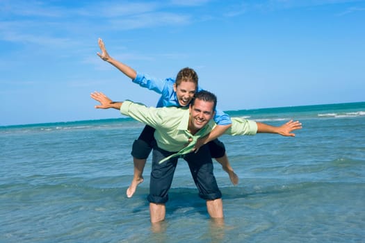 Mid adult woman riding piggyback on a mid adult man with their arms outstretched on the beach