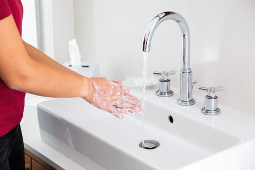 Woman lathering hands under running water in white bathroom. Person cleaning skin with soap in washbasin. Female with foam on arms under flowing water.