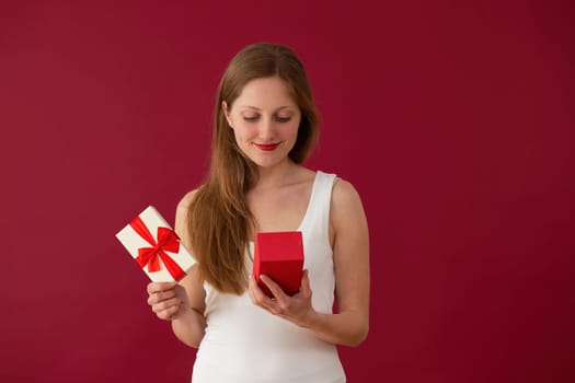 Blonde girl looking to the present on red background. Smiling woman holding valentine s gift. Female with lipstick with box with ribbon on burgundy backdrop smiling over a present.