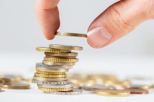 Fingers putting one euro on pile of coins from close up. Concept of saving money for future. Adding cash on pile with white background.