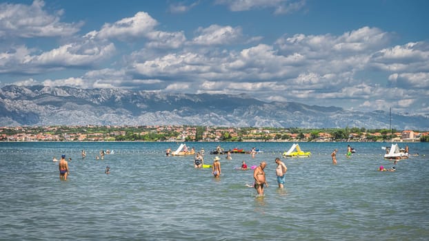 Nin, Croatia, July 2019 People relaxing and having fun in water of Adriatic Sea, Zdrijac beach with mountain range of Dinaric Alps in the background