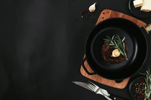 Cooked ribeye steak on cast-iron pan and board with pepper, rosemary, salt, garlic, knife, fork, butter on black background