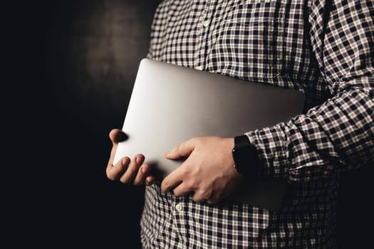 Man wearing casual clothes holds closed laptop, black blurred background. High quality photo