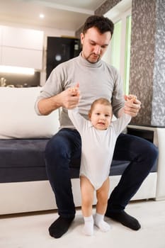 Young father assisting walking his infant son teaching to walk at home. Daddy helps son attempting to walk