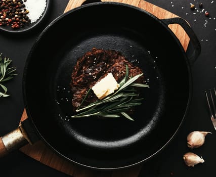 Cooked ribeye steak on cast-iron pan and board with pepper, rosemary, salt, garlic, knife, fork, butter on black background