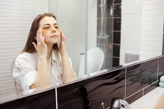 Caucasian woman applying hydrogel patches on under eye area, looking at mirror. Happy young lady enjoying morning anti age anti wrinkle routine in bathroom.