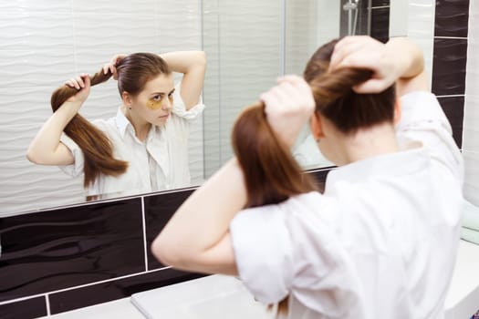 Young woman with hydrogel patches under eyes makes ponytail looking into the mirror in bathroom.